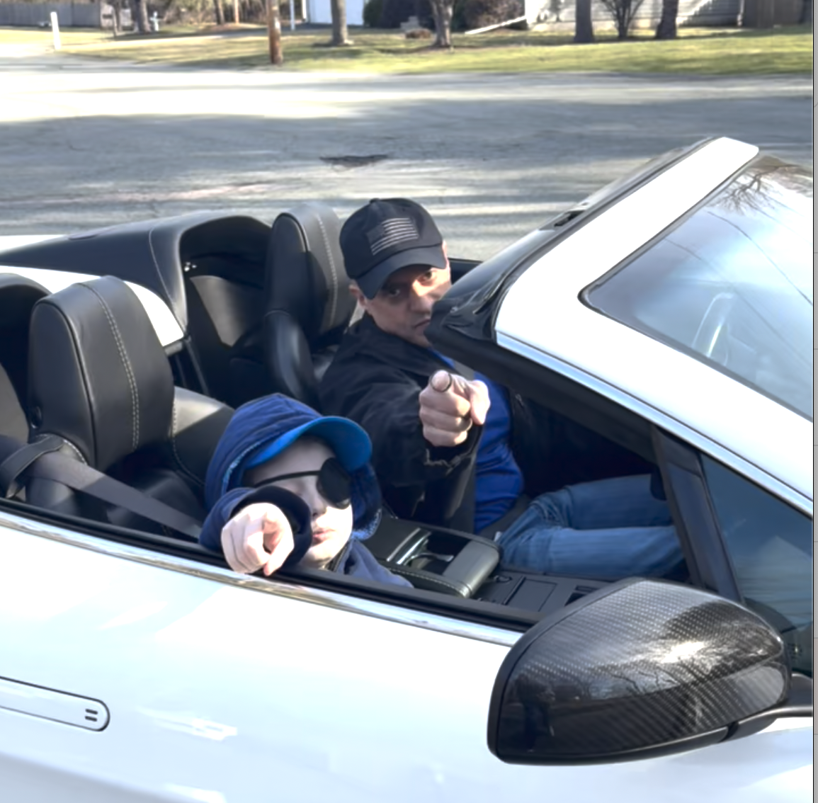 A young boy and middle age man point to the camera in a white convertable